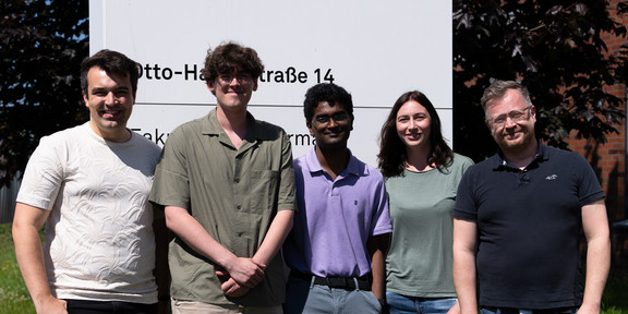 Five smiling people in front of a sign of TU Dortmund