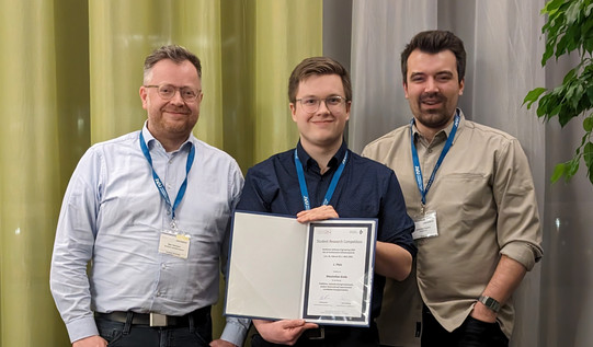 Three smiling persons next to each other. The person in the middle is holding a certificate.