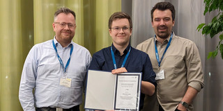 Three smiling persons. The person in the middle is holding a certificate.