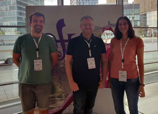 Three smiling researchers in front of the ASE conference photo wall in Luxembourg.